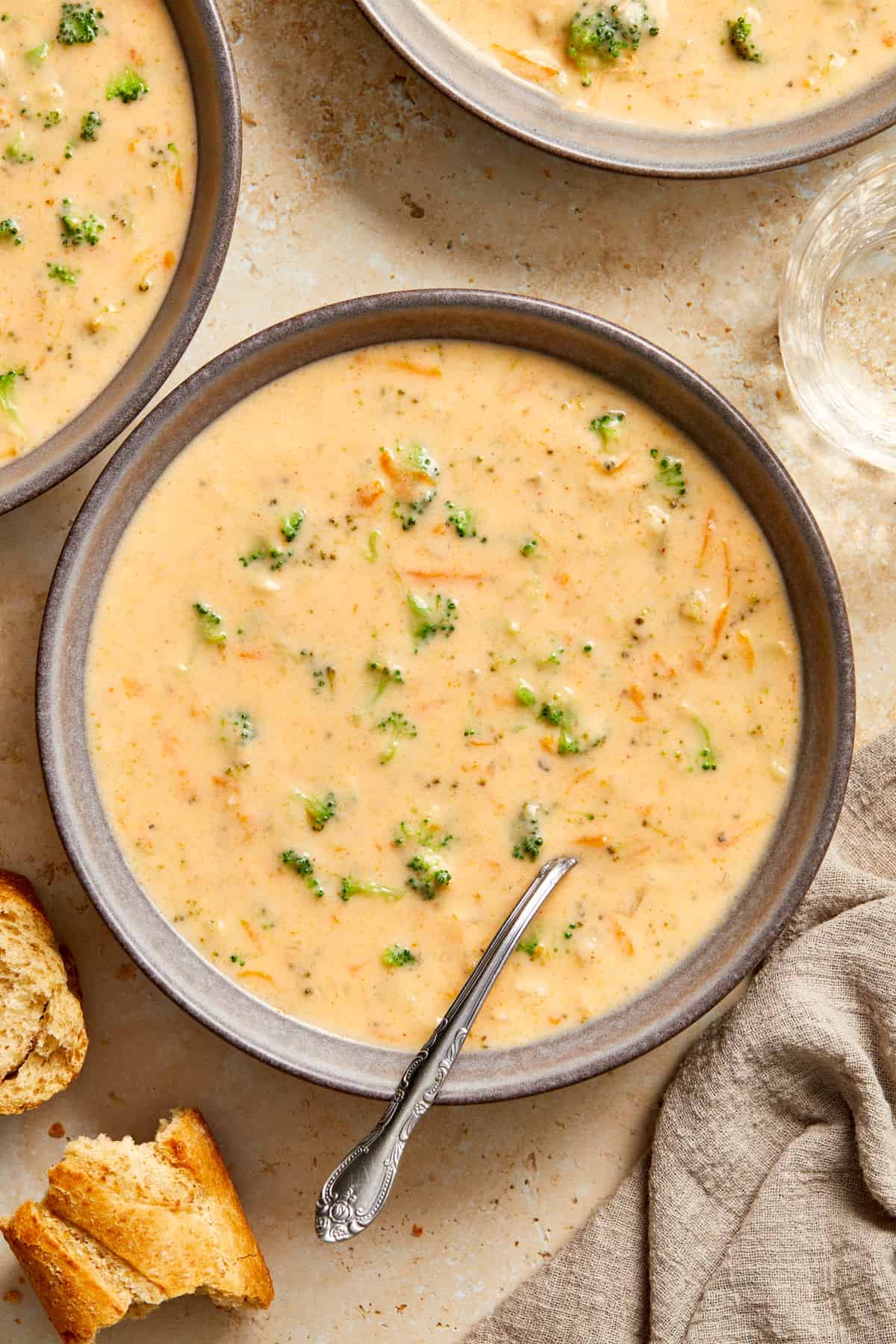 Gray bowl with broccoli cheddar soup with spoon along with a chunk of bread beside it.