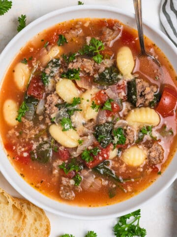 Overhead view of white bowl with sausage gnocchi soup with spoon and sliced bread and linen surrounding.