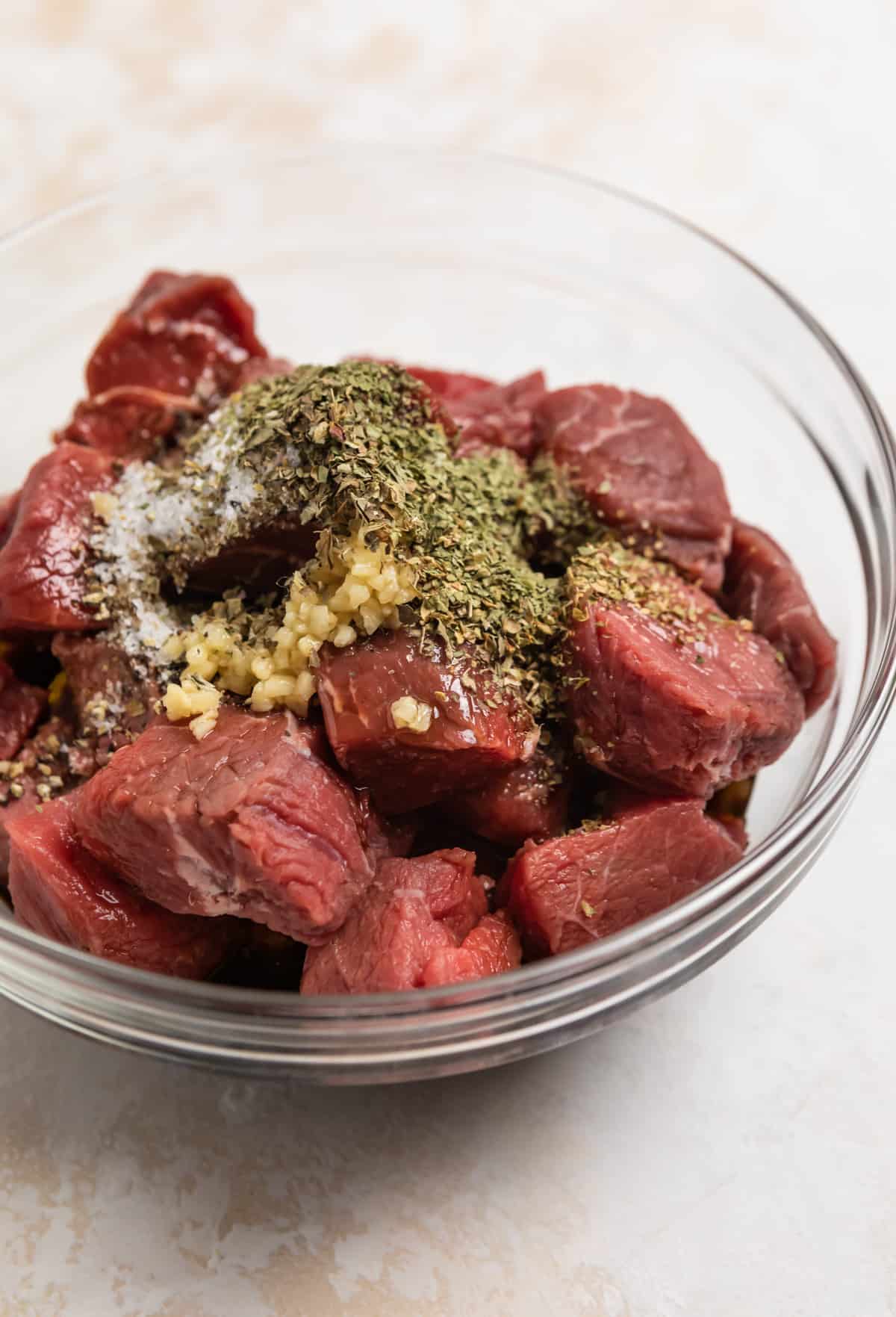 Steack cubes in glass bowl with garlic, herbs and marinade ingredients.