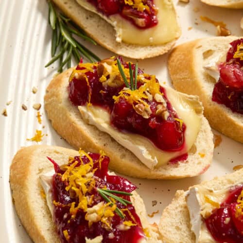 Cranberry topped brie crostini on plate topped with fresh rosemary and orange zest.