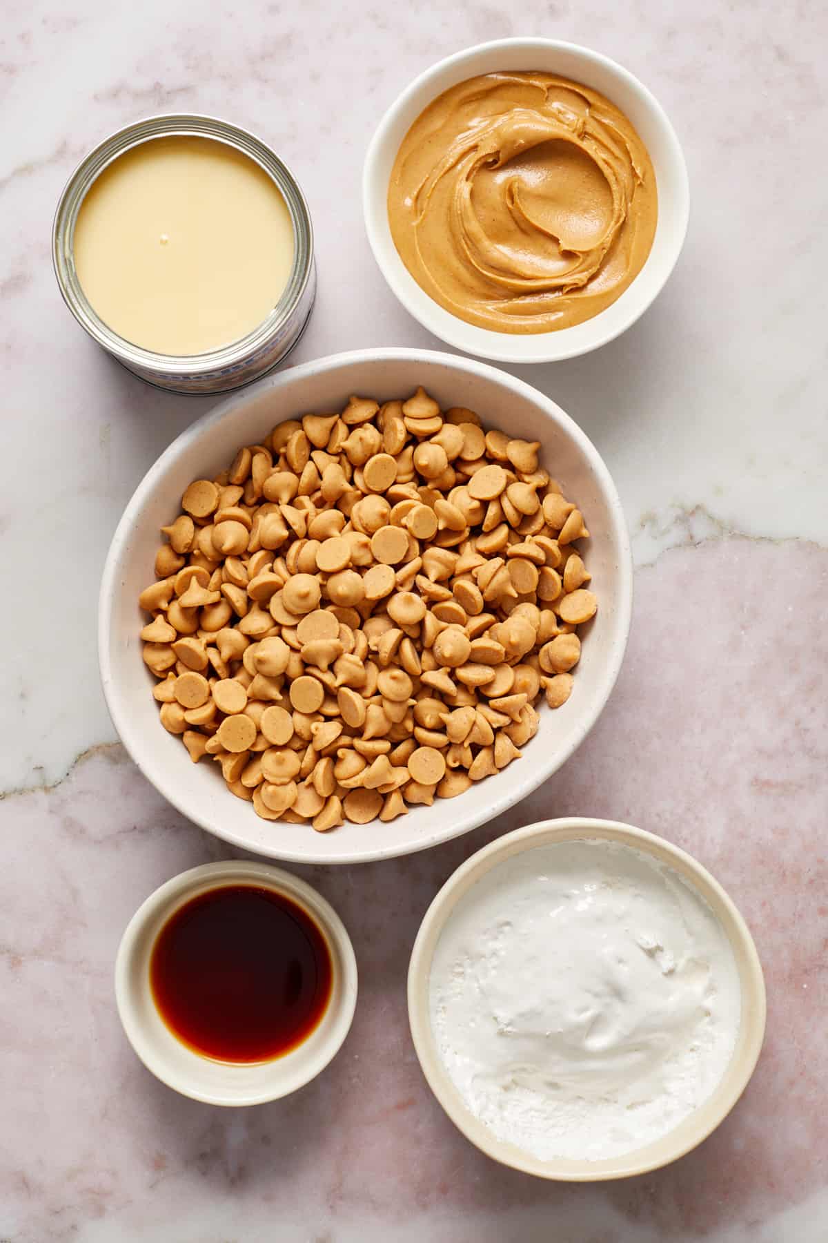 Peanut butter chips, condensed milk, marshmallow cream and vanilla in bowls on surface.