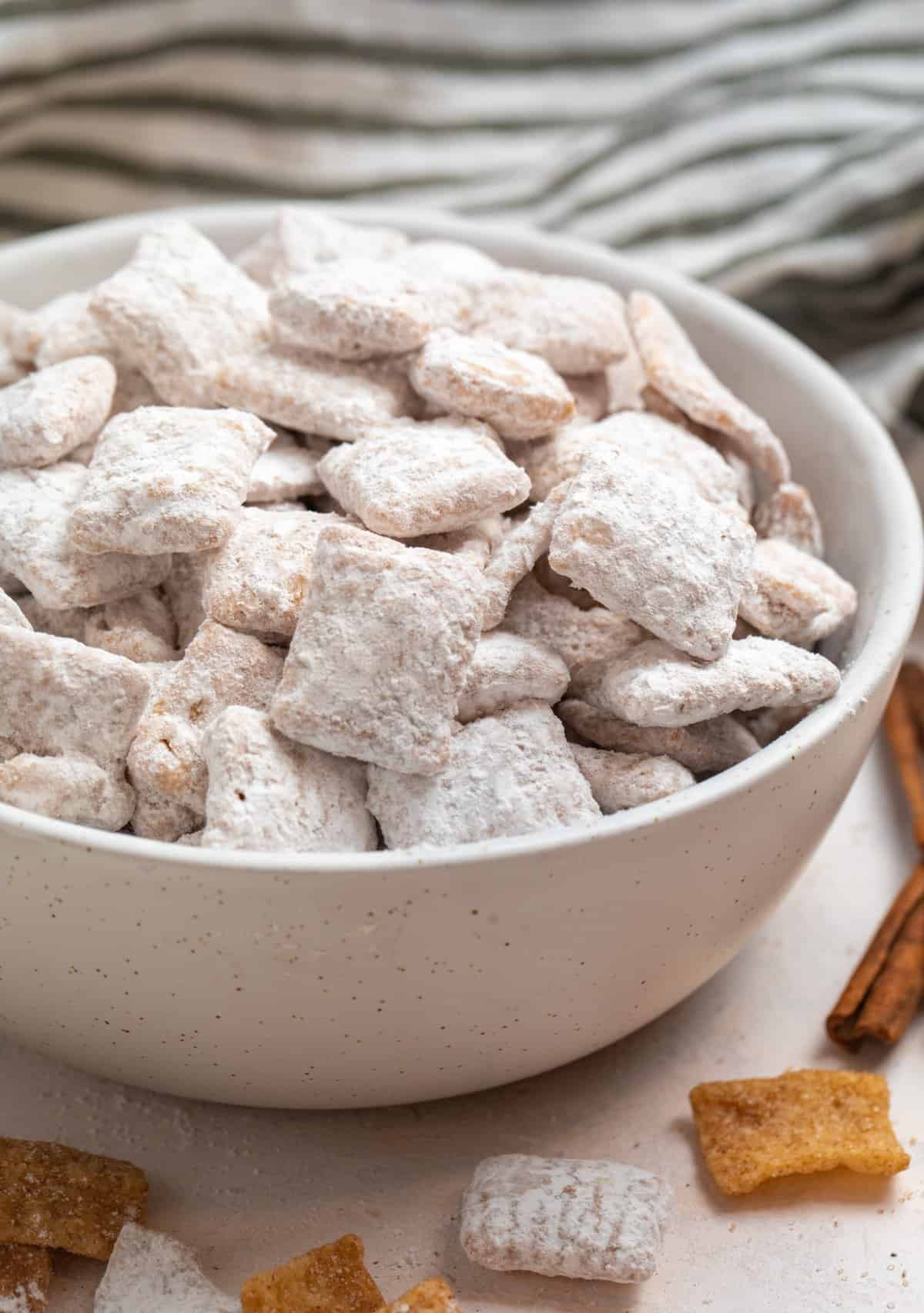Snickerdoodle puppy chow in bowl with cinnamon stick and cereal beside it.