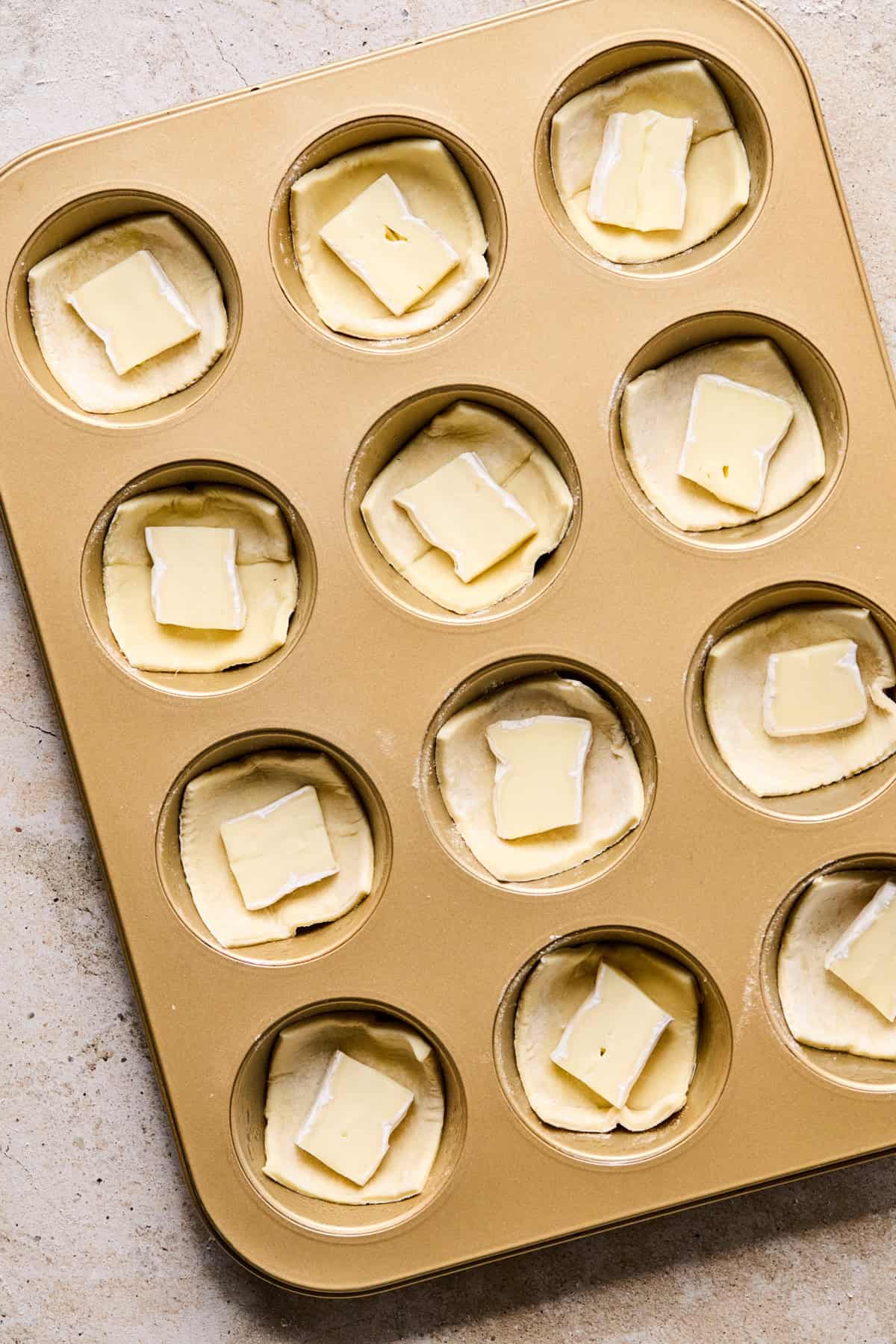 Muffin tin with puff pastry squares filled with piece of brie cheese.