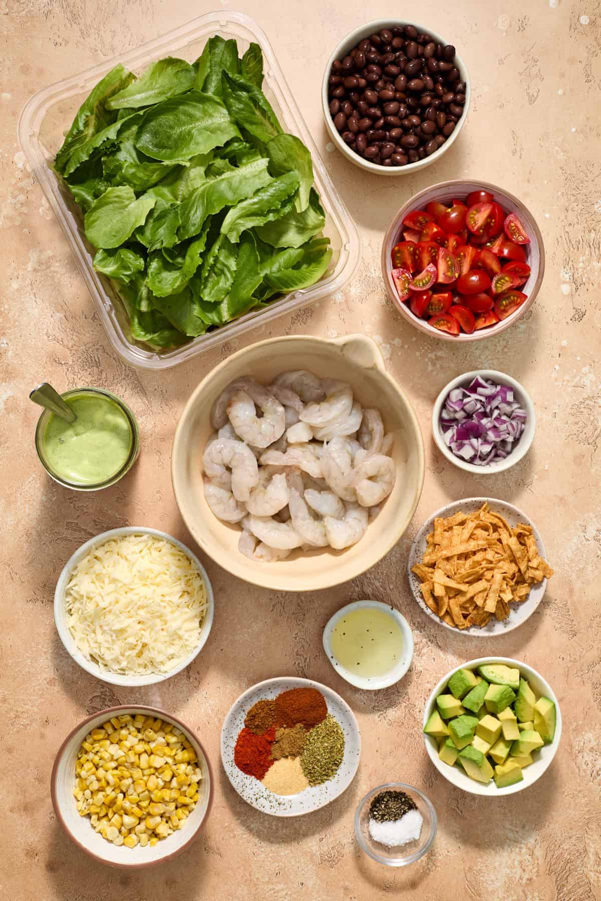 Uncooked shrimp, tomatoes, lettuce, beans and other taco salad ingredients arranged on surface.