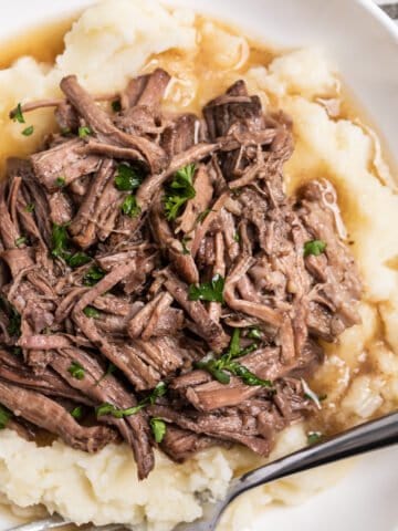 White plate with crock pot shredded beef over mashed potatoes.