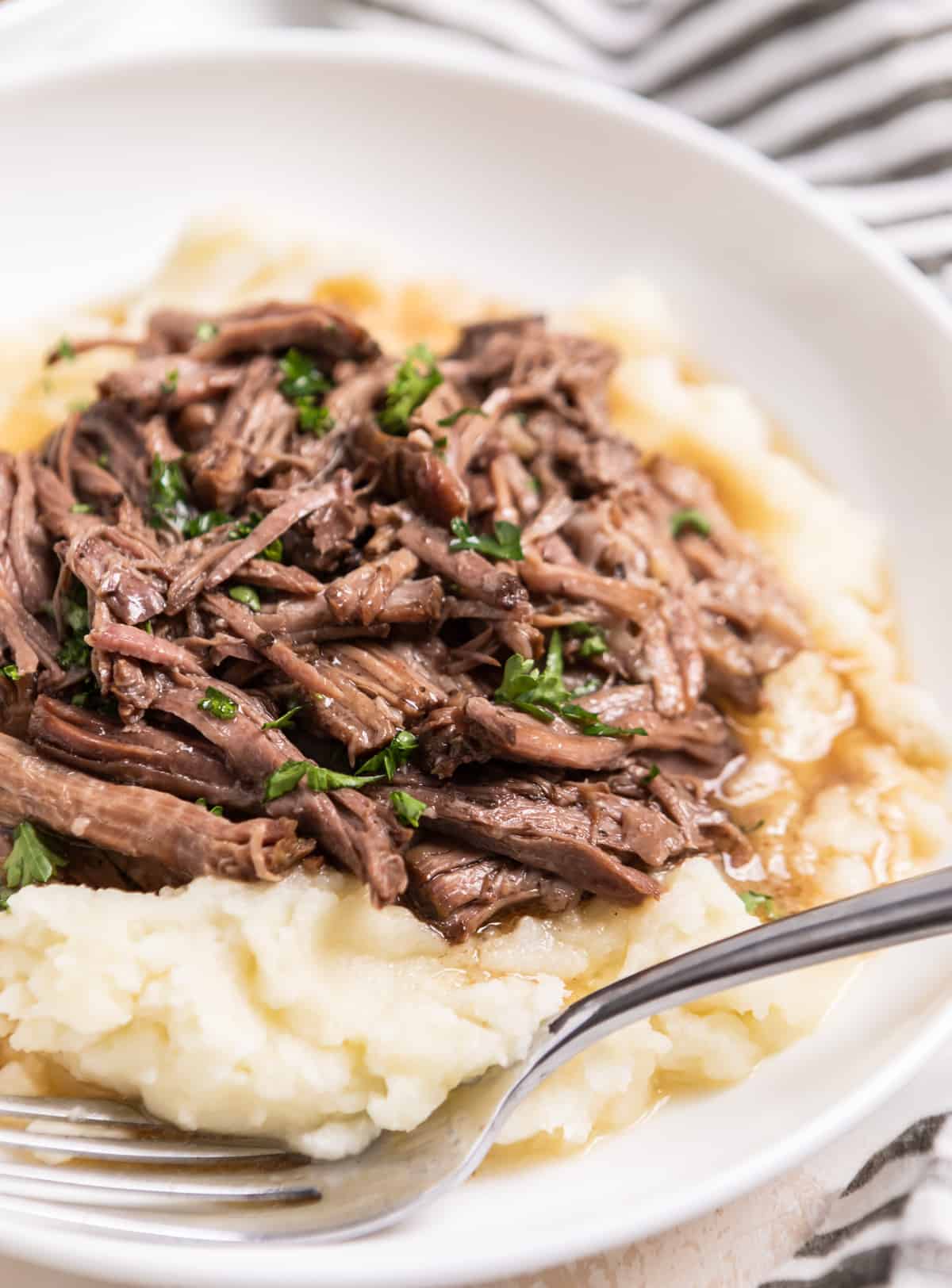 White plate with slow cooked shredded beef on mashed potatoes with chopped parsley.
