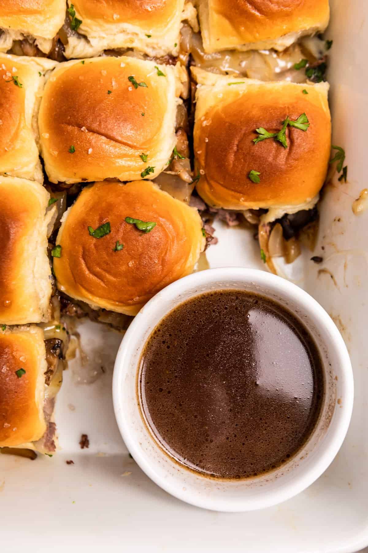 Overhead view of au jus in bowl with roast beef sliders surrounding it.
