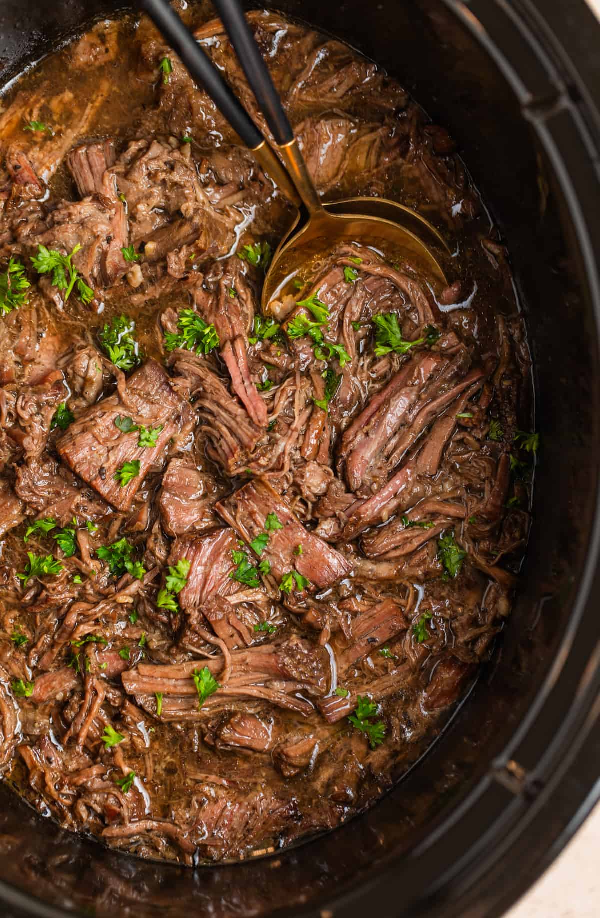 Slow cooker shredded beef in crockpot with serving utensils and chopped parsely.