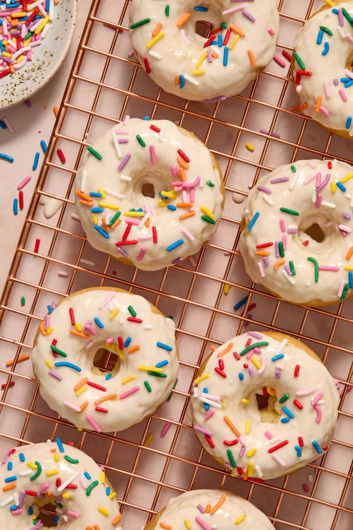 Baked vanilla donuts with icing glaze and rainbow sprinkles on cooling rack.