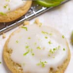 Key lime iced cookie on counter with crumbs, lime and other cookies surrounding it.