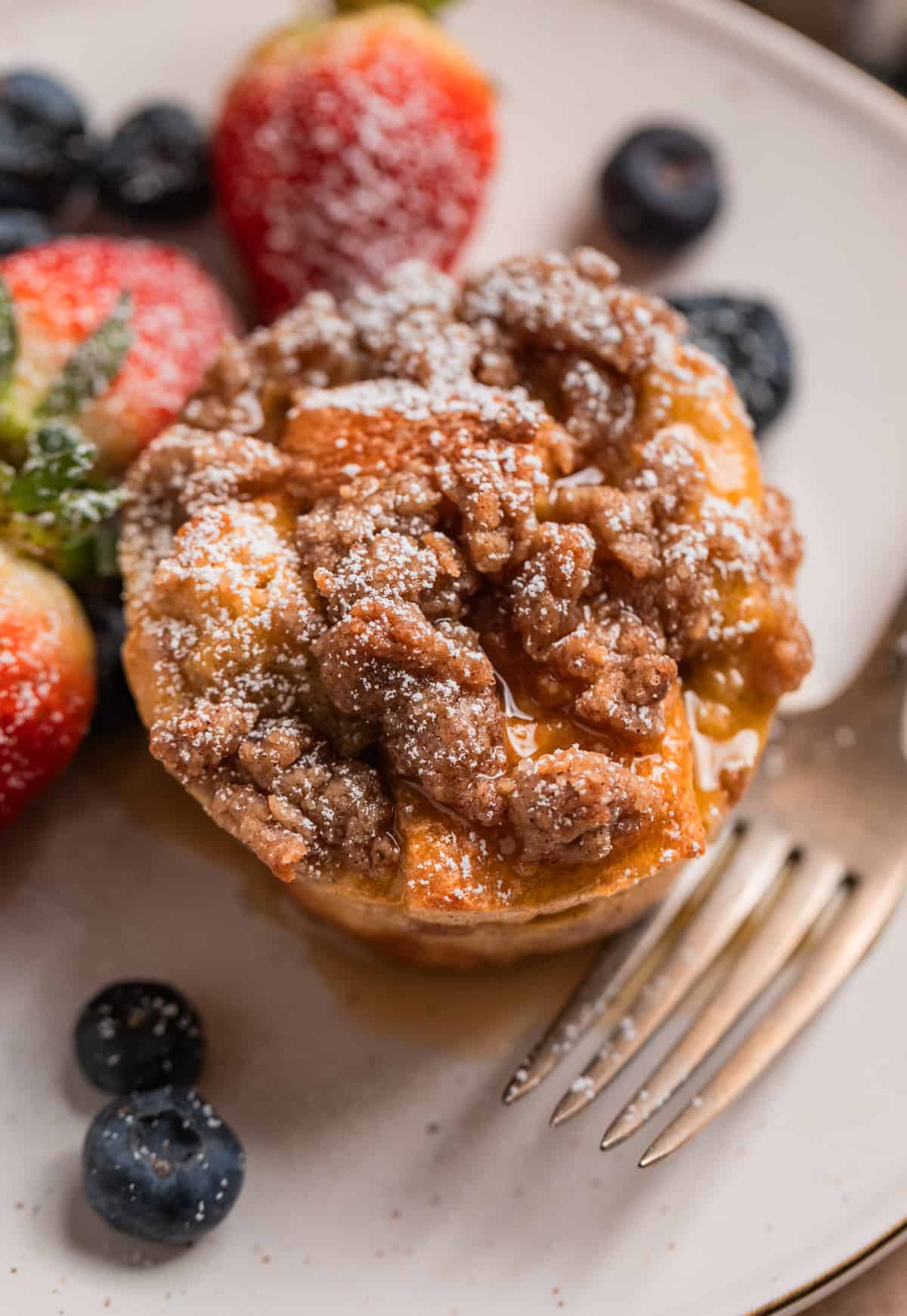 French toast muffin with streusel topping, maple syrup and powdered sugar on plate with berries and fork.