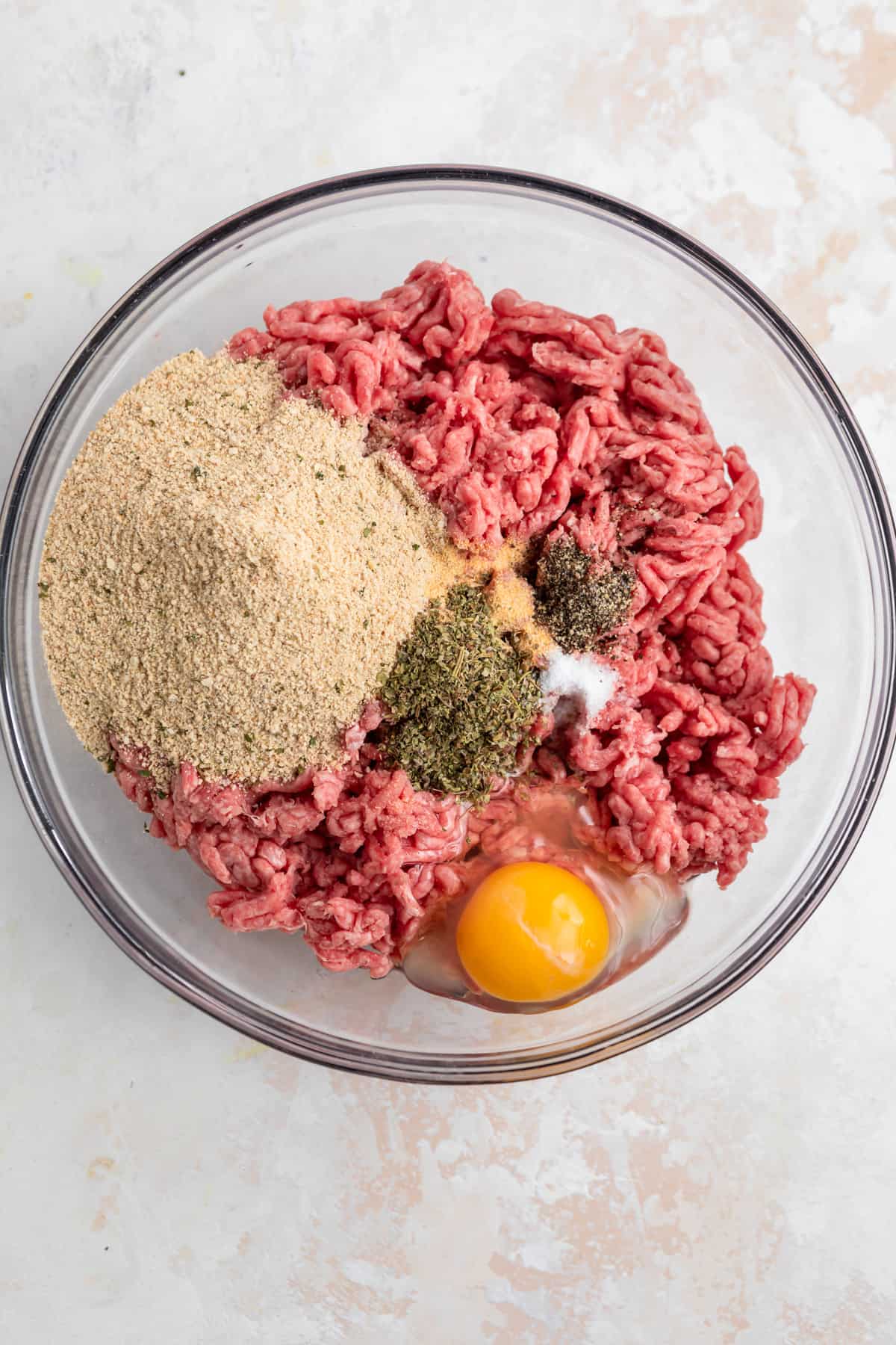 Glass bowl with ground beef, breadcrumbs, egg, seasoning and salt and pepper.