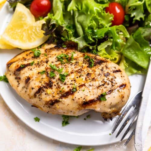 Grilled lemon pepper chicken on white plate with lemon slices and salad.