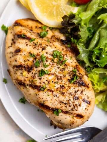 Lemon pepper chicken on plate with salad, lemon wedges and fork and knife.