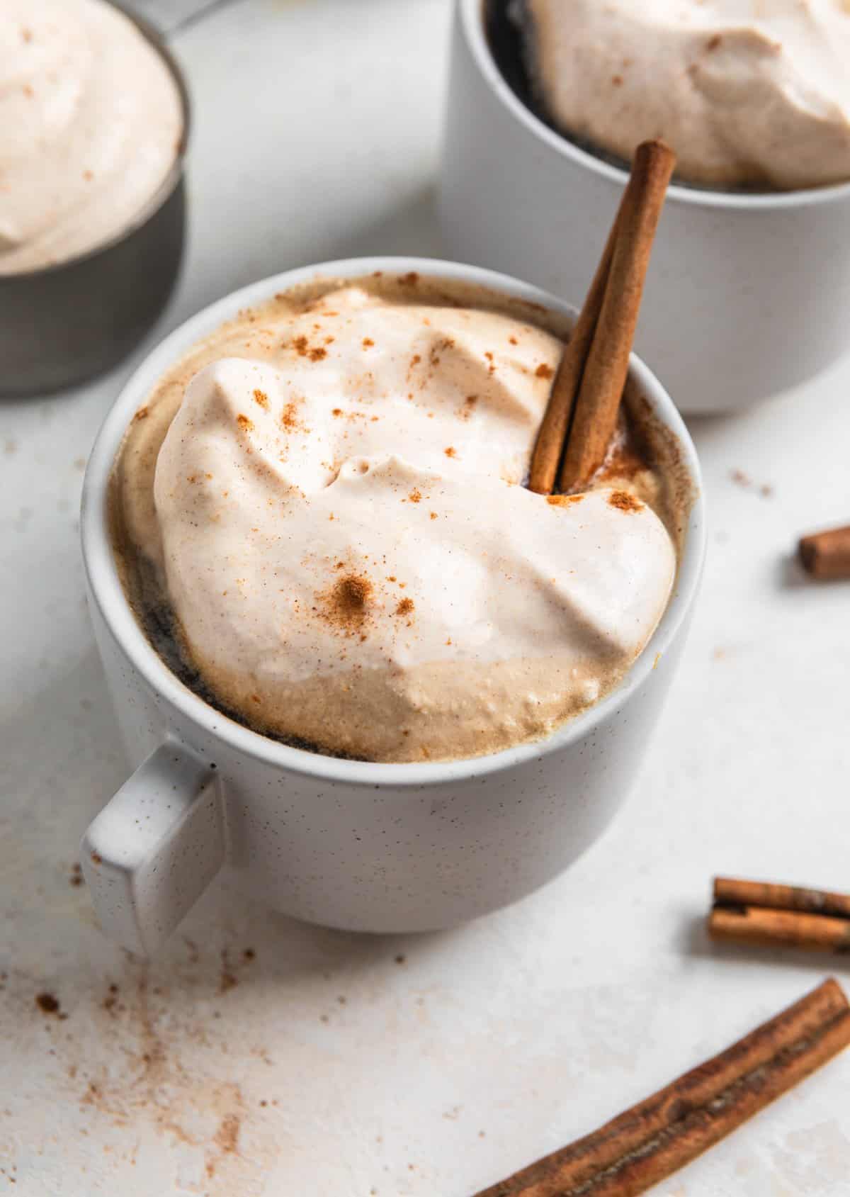 Coffee cup with pumpkin whipped cream dollop on top and a cinnamon stick.