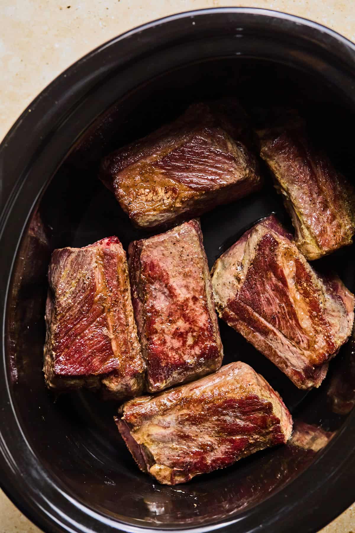 Crockpot with seared short ribs arranged in the bottom of the pot.