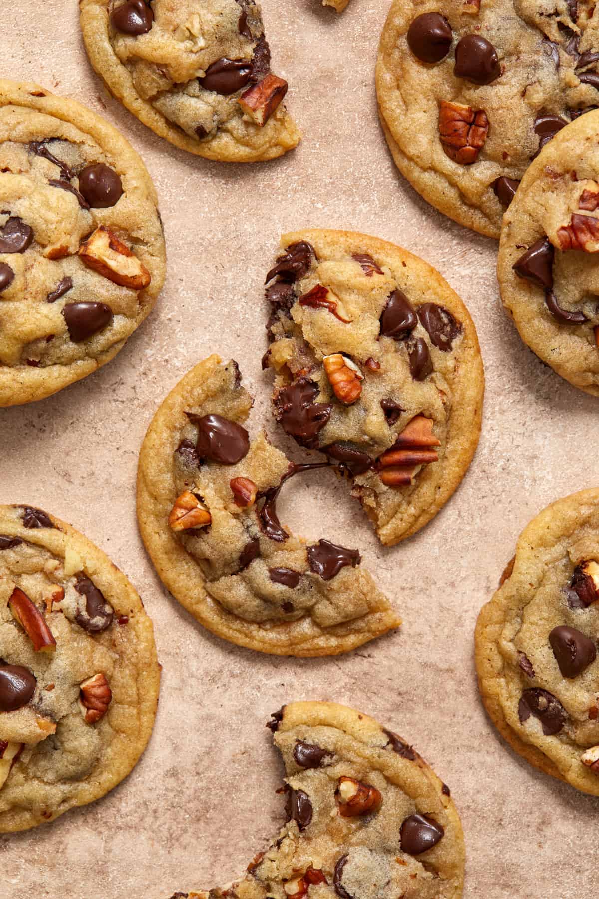 Chocolate chip cookies with pecans on surface with focus on center cookie broken in half with melted chocolate string connecting the pieces.