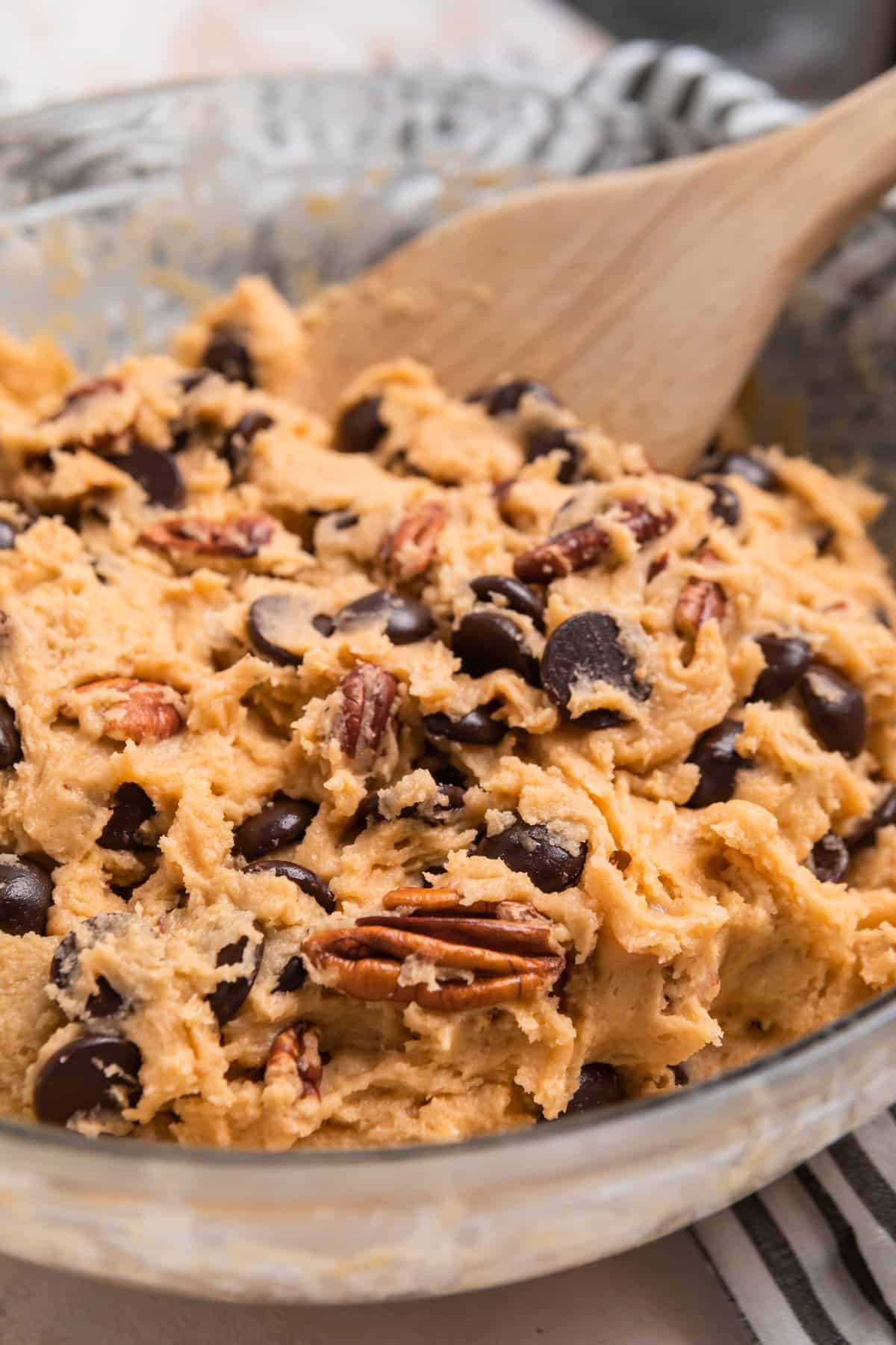 Chocolate chip pecan cookie dough in glass mixing bowl with wood spoon.