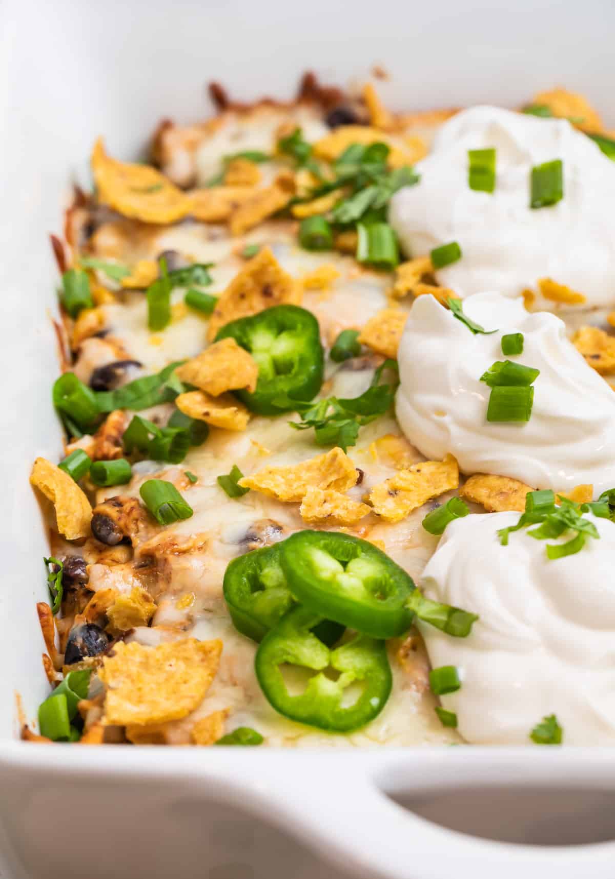 Chicken taco casserole in casserole dish topped with Fritos, jalapeño, cilantro and sour cream.