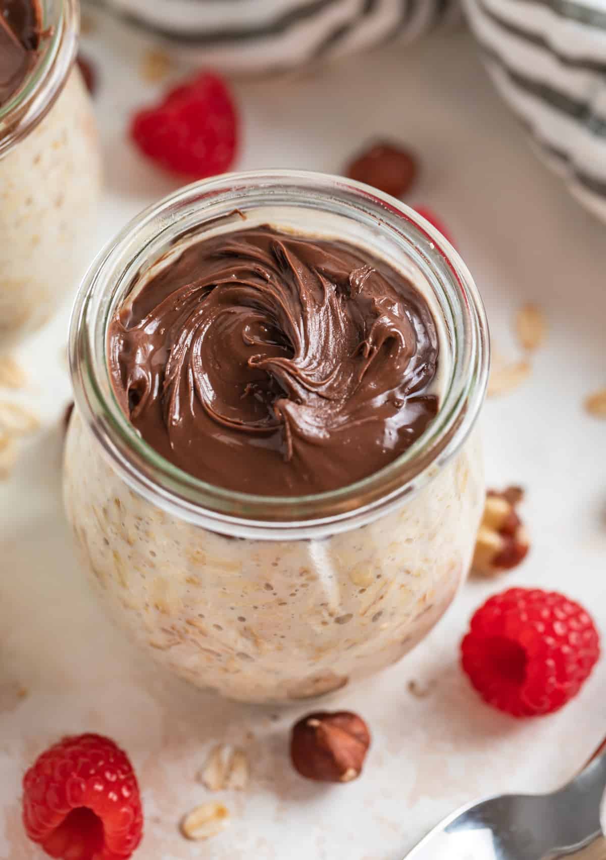 Jar of overnight oatmeal with nutella swirled on top and raspberries and hazelnuts beside.