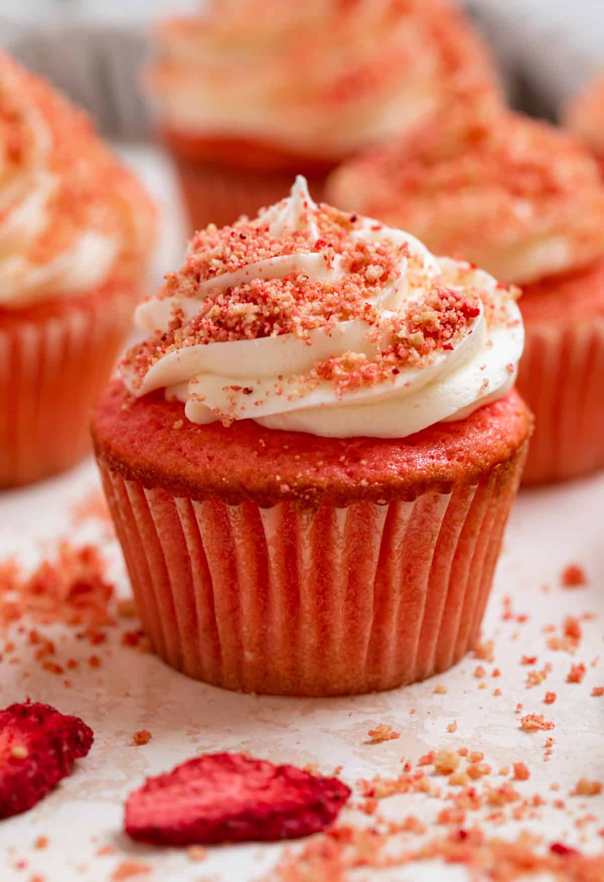 Strawberry cupcakes with buttercream frosting and strawberry crunch topping lined on counter with freeze dried strawberries and crumbs.