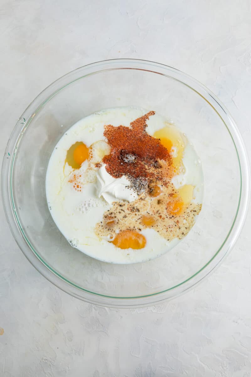 Eggs, seasoning, milk and sour cream in glass mixing bowl.