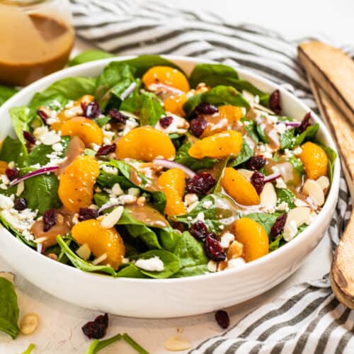 White salad bowl with spinach, cranberries, almonds, goat cheese, oranges and balsamic dressing drizzled on top.
