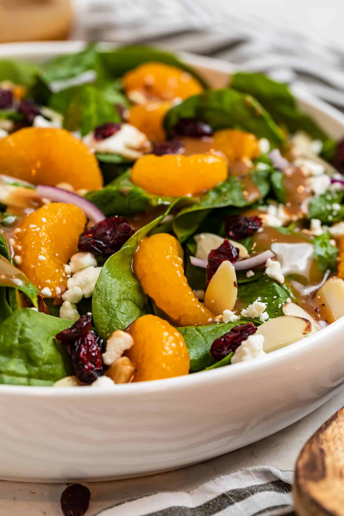 White bowl with spinach, mandarin oranges, almonds and more with wood spoon beside it.