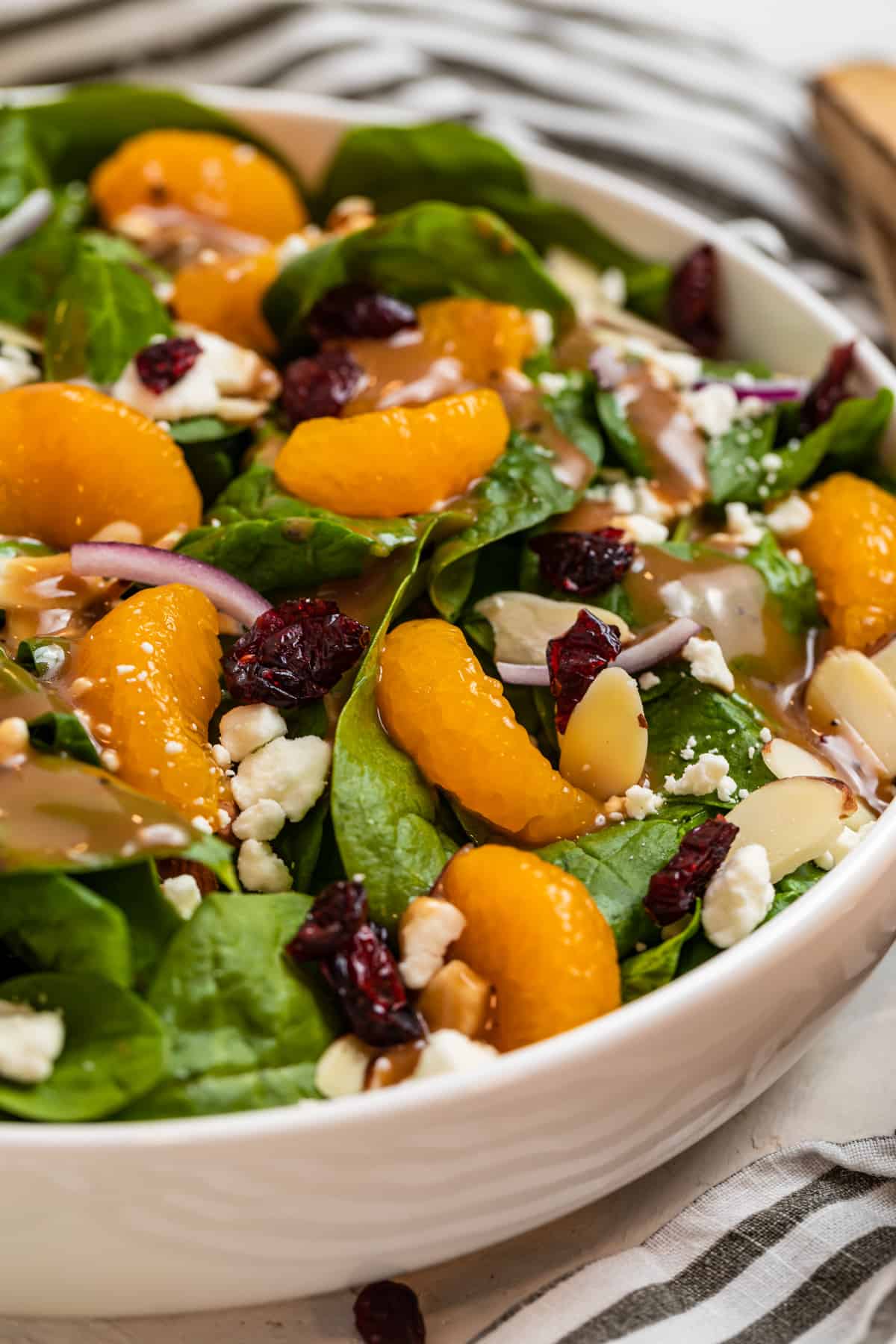 White bowl with spinach mandarin salad topped with goat cheese, cranberries, almonds and honey balsamic dressing.