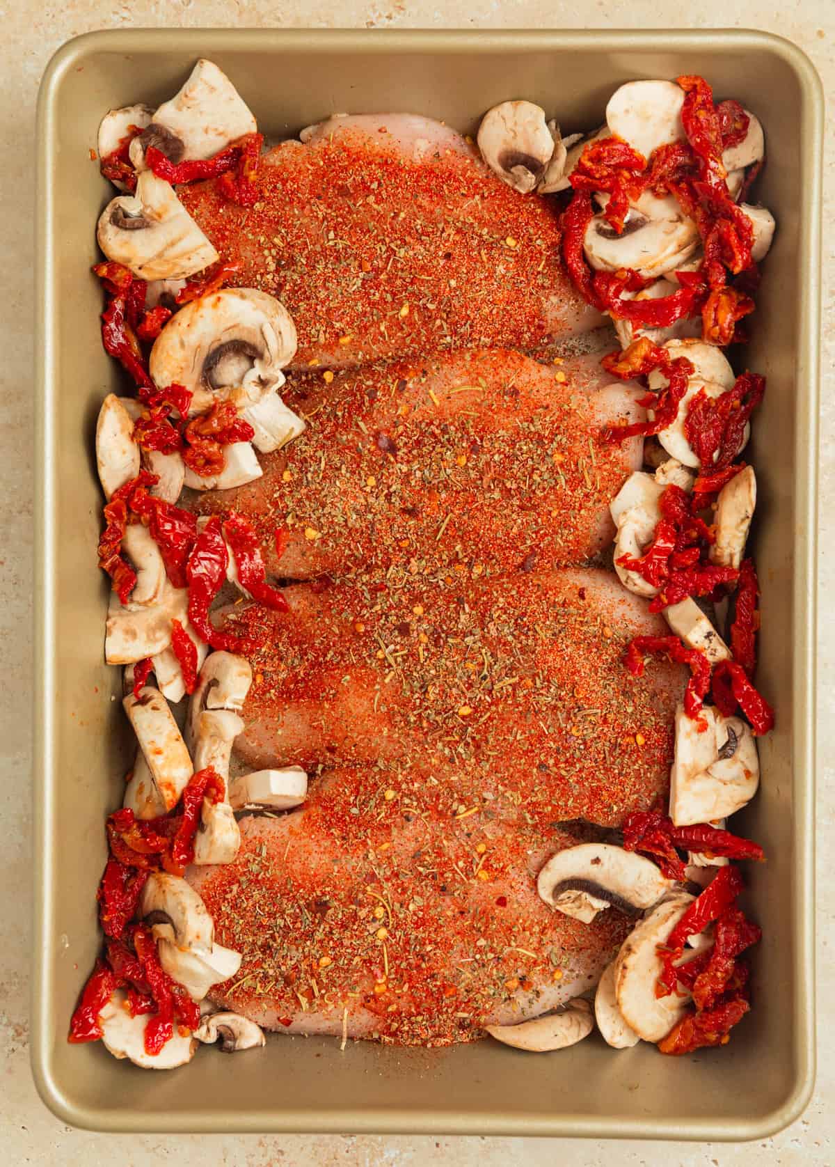 Baking pan with seasoned chicken with mushrooms and sun-dried tomatoes lining the edges of the pan.