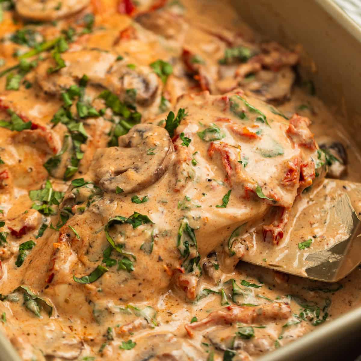 Serving spatula picking up piece of creamy baked tuscan chicken topped with mushrooms, parsley, etc.