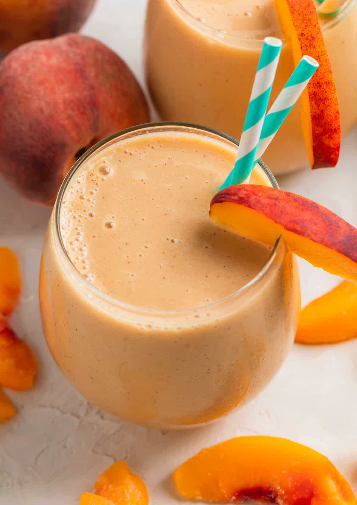 Peach banana smoothie in glass with two straws and peach slice garnish.