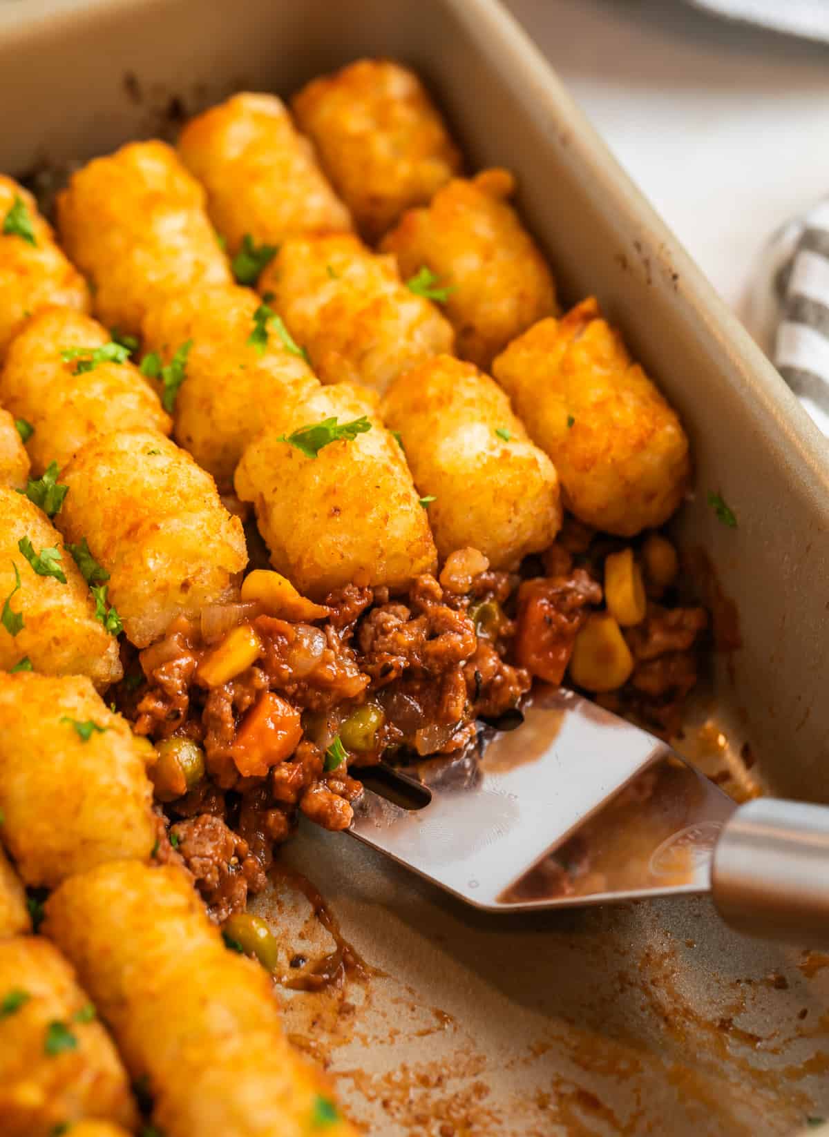 Tater tot topped beef mixture in baking pan with spatula scooping into the dish.