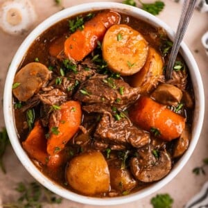 White bowl with crockpot beef stew with mushrooms, carrots and potatoes topped with chopped parsley.