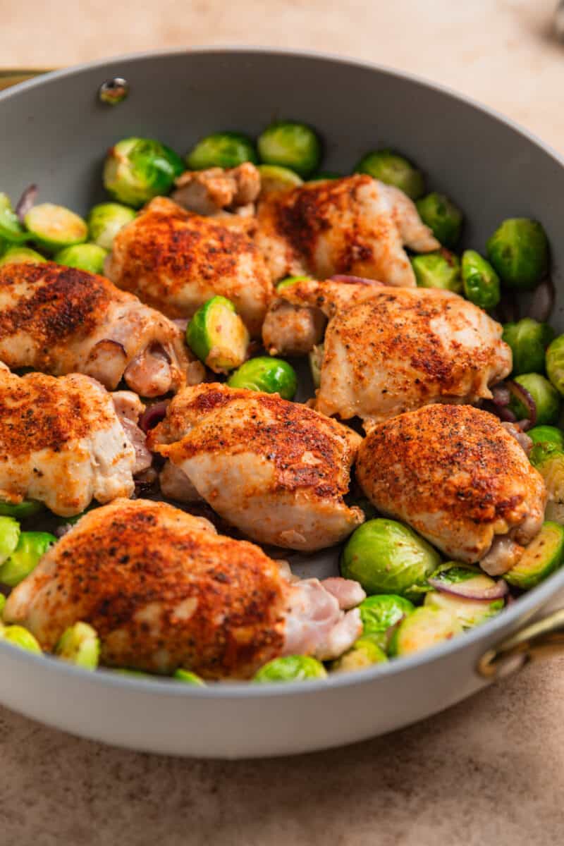 Chicken thighs and Brussel sprouts being cooked in gray skillet.