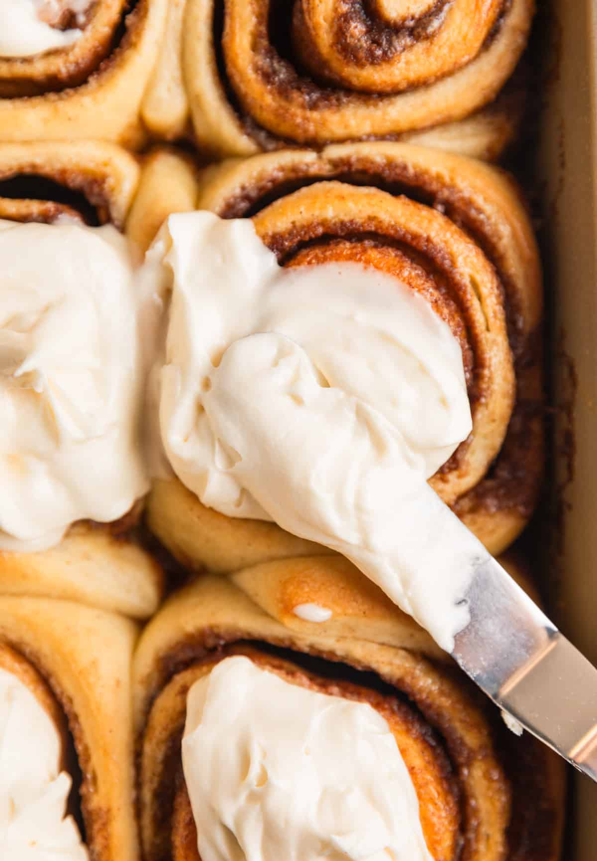Cinnamon rolls in baking pan with icing being spread over top with offset spatula.