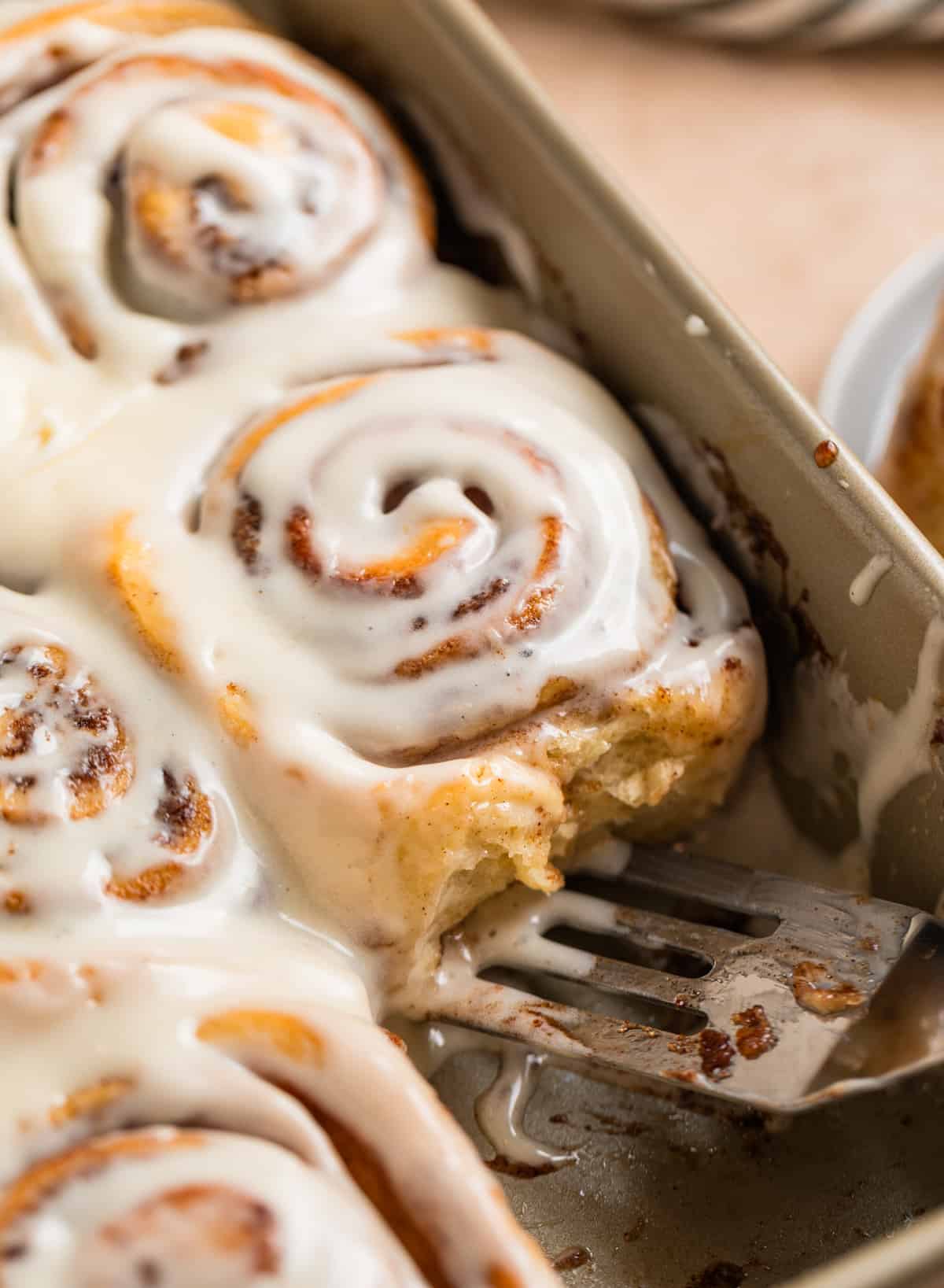 Iced buttermilk cinnamon rolls in pan with spatula scooping into the pan.