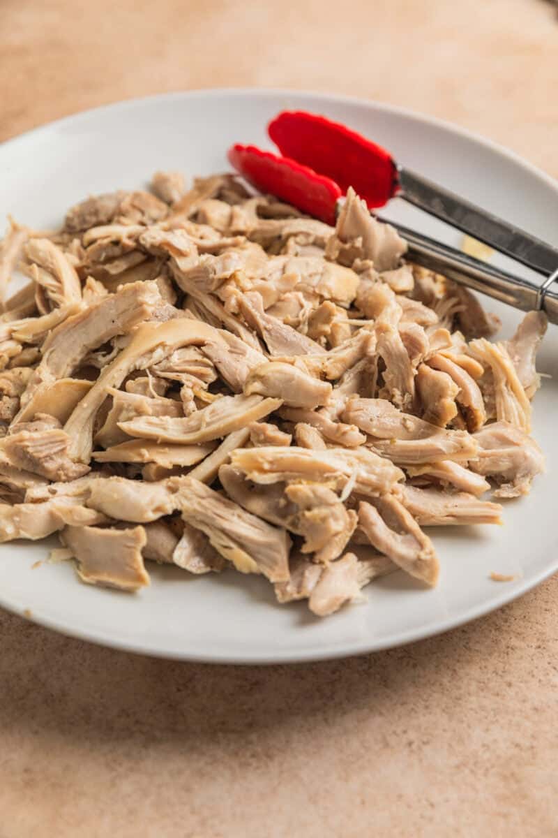 Shredded chicken thighs on white plate with tongs.