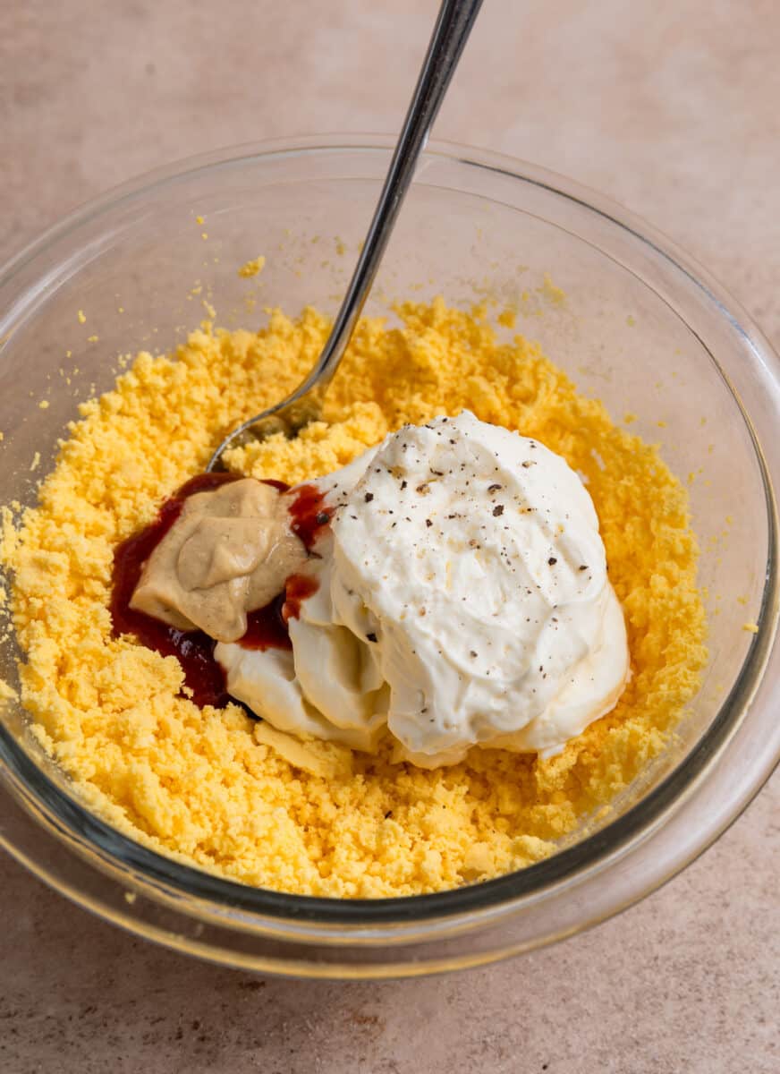 Mashed egg yolk, mayonnaise, dijon mustard and other ingredients in glass bowl with spoon. 