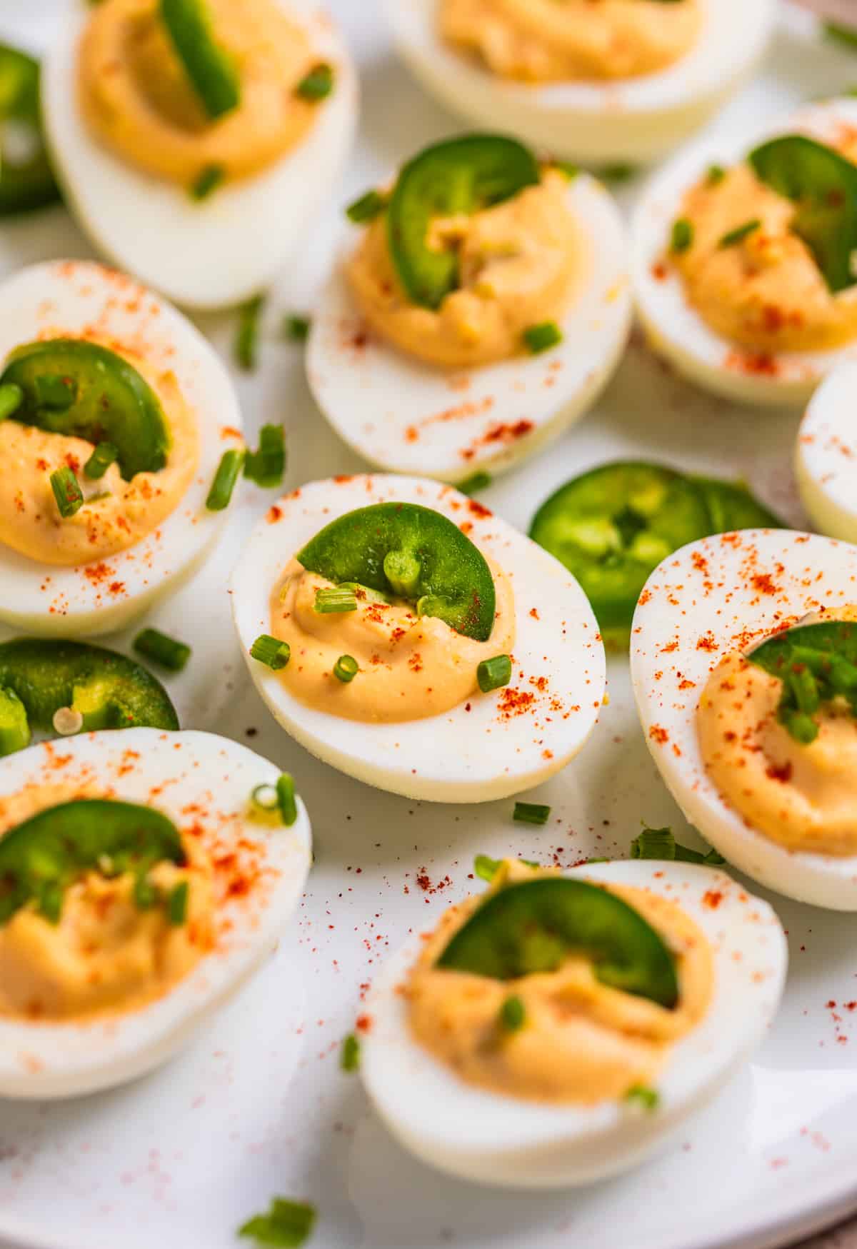 White dish with spicy jalapeño deviled eggs garnished with chives, paprika and sliced jalapeño.