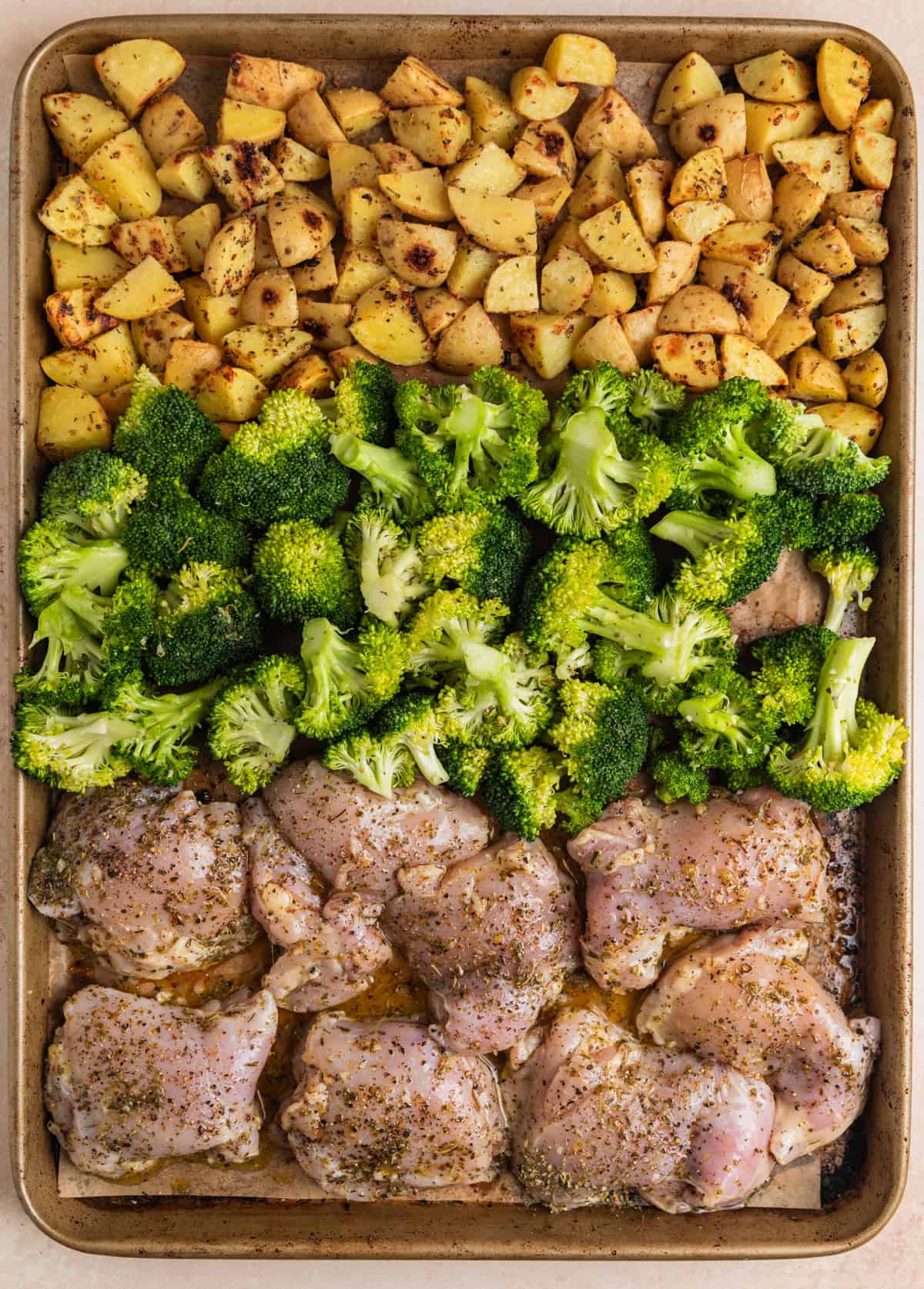 Uncooked chicken thighs and broccoli added to sheet pan with potatoes already partially roasted.