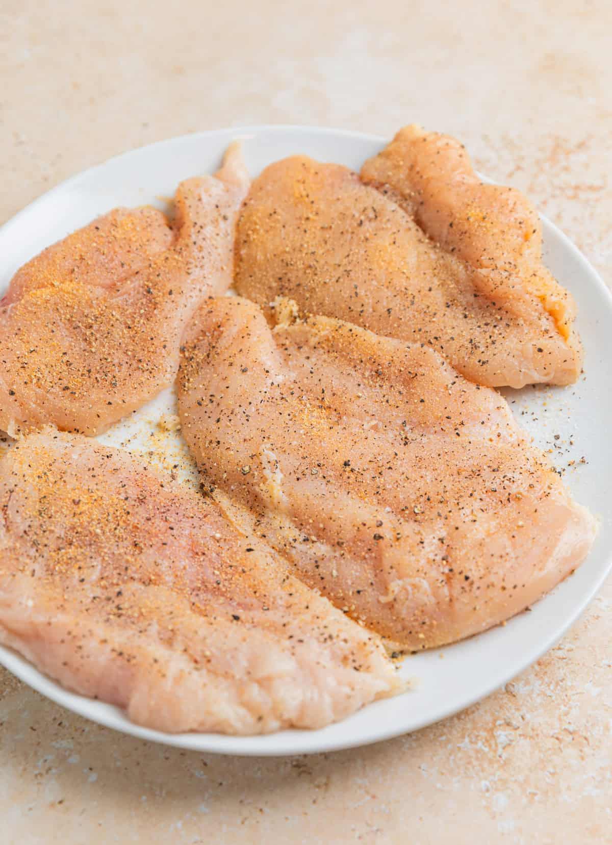 Seasoned thin chicken breasts on white plate before cooking.