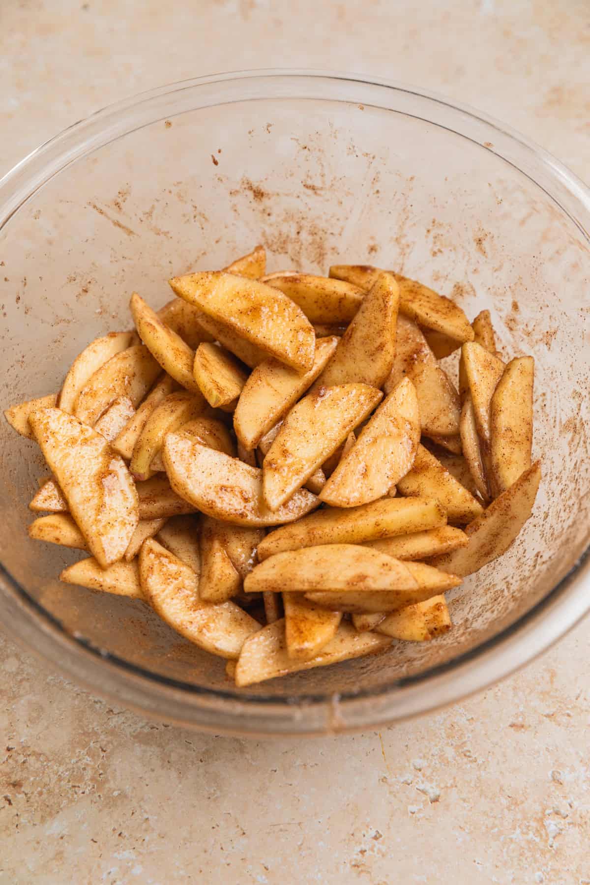 Apple slices tossed in cinnamon and sugar in glass bowl.