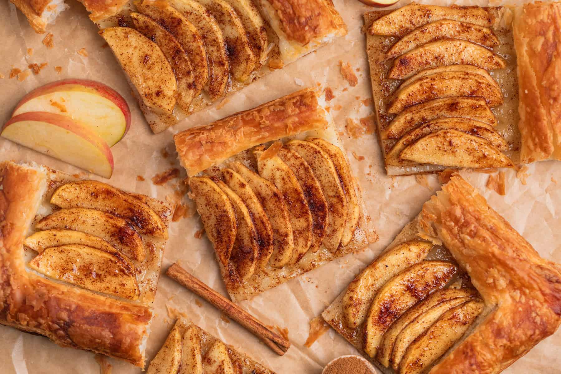 Puff pastry apple tarts arranged on parchment with apple slices and cinnamon sticks.