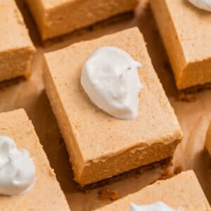 Pumpkin cheesecake bars arranged on brown parchment topped with a dollop of whipped cream.