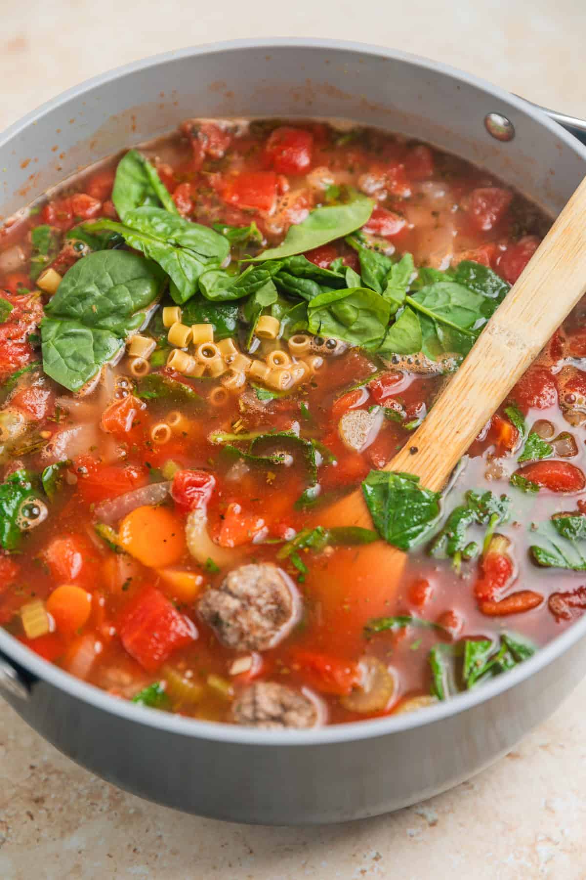 Meatballs, pasta and spinach added to soup in pot with wooden spoon propped into it.