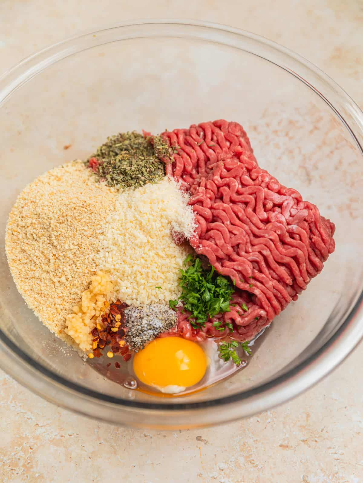 Ground sirloin, breadcrumbs, egg, parsley and other meatball ingredients in glass mixing bowl.