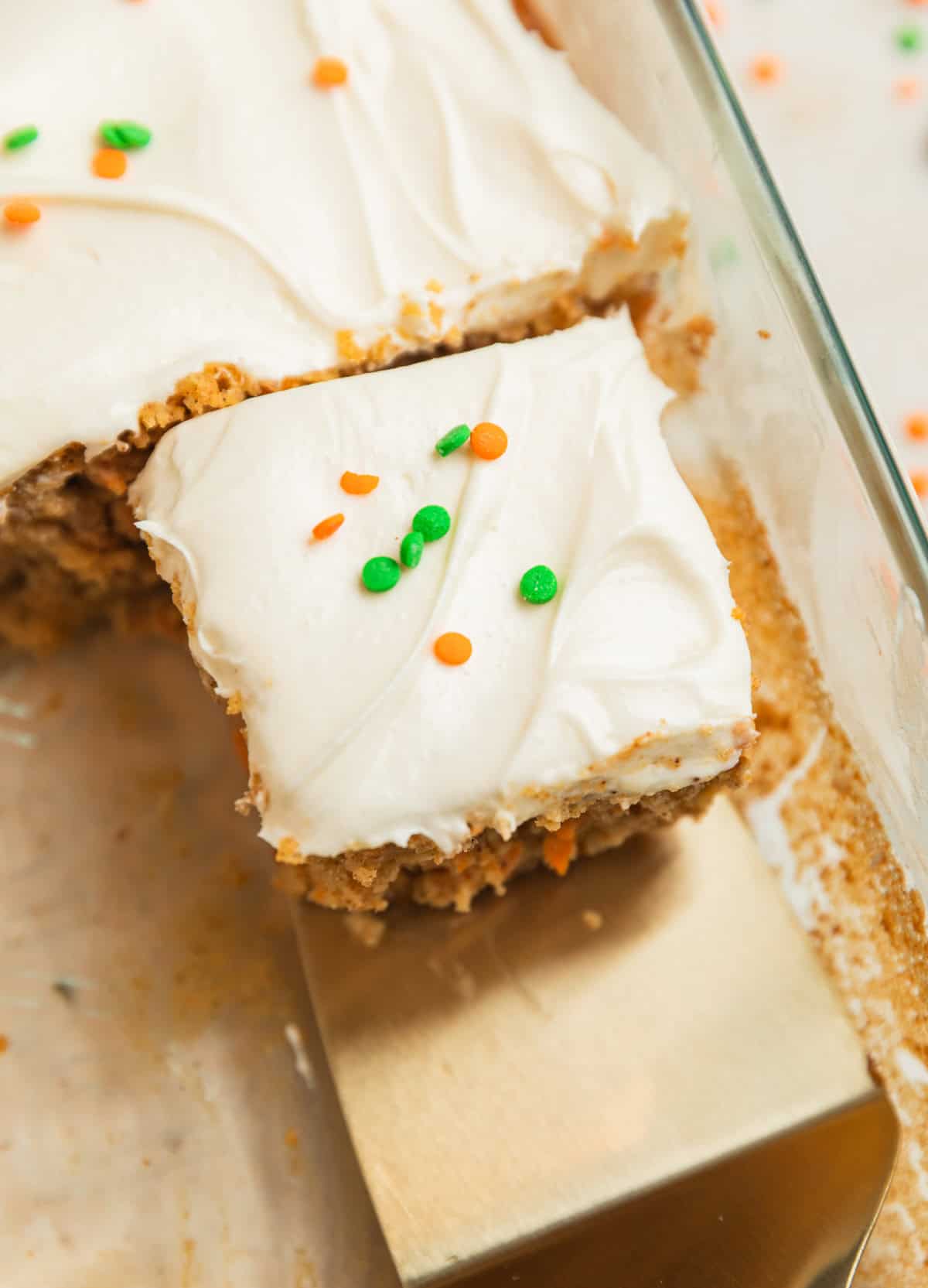 Slice of carrot cake in baking pan topped with cream cheese frosting and orange and green sprinkles.