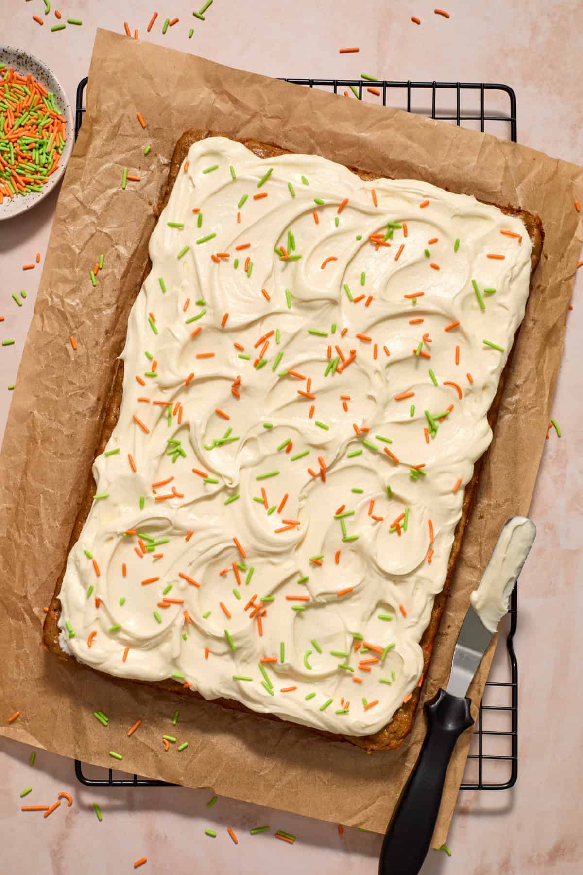 Cream cheese frosted carrot cake on parchment paper with orange and green sprinkles on top.