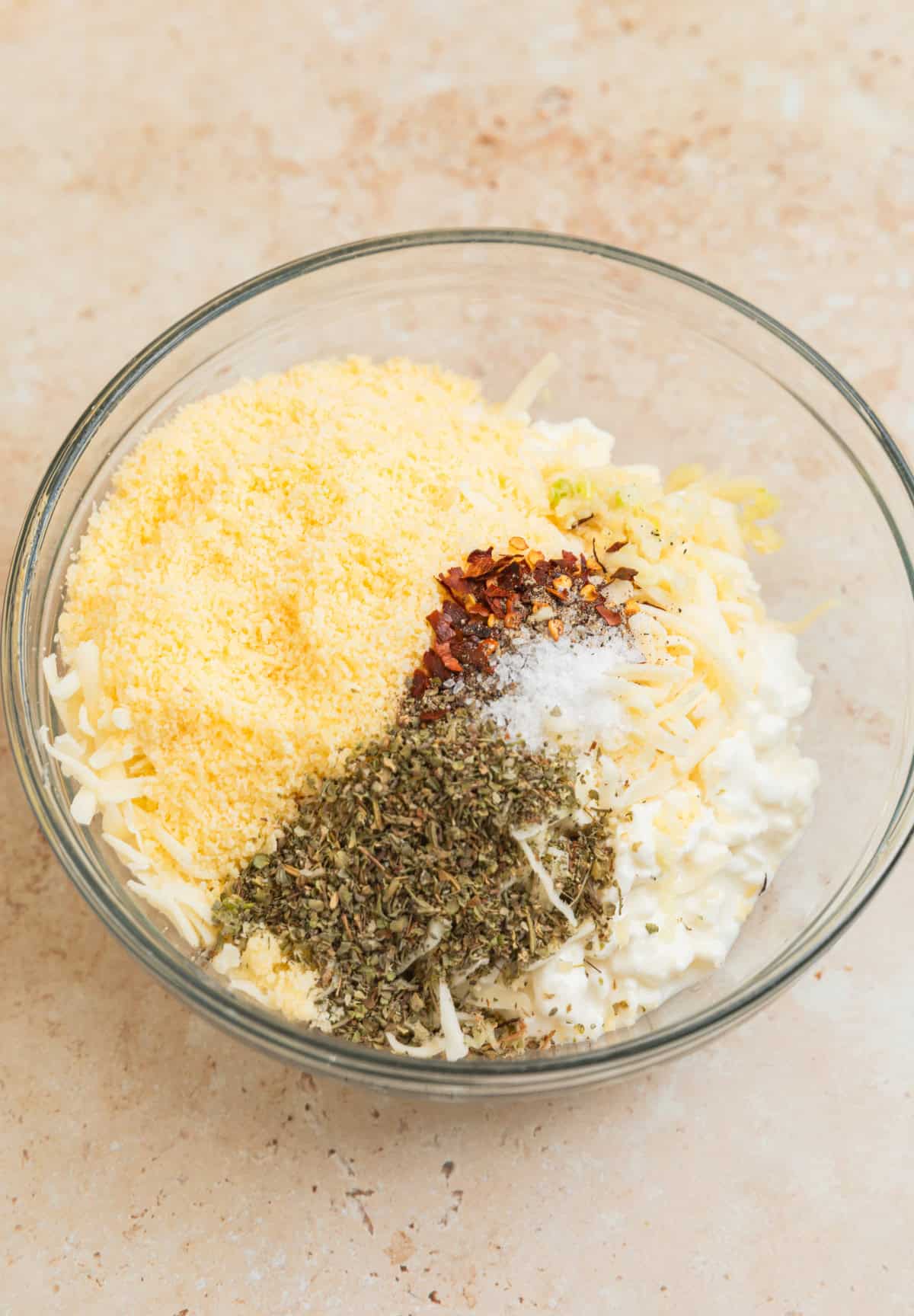 Cottage cheese, parmesan cheese, spices and other ingredients in glass bowl before stirring.